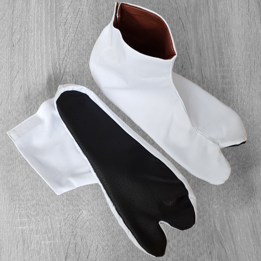 A pair of white split-toe tabi shoes with brown leather soles, designed for aikido and other martial arts, displayed on a wooden floor.