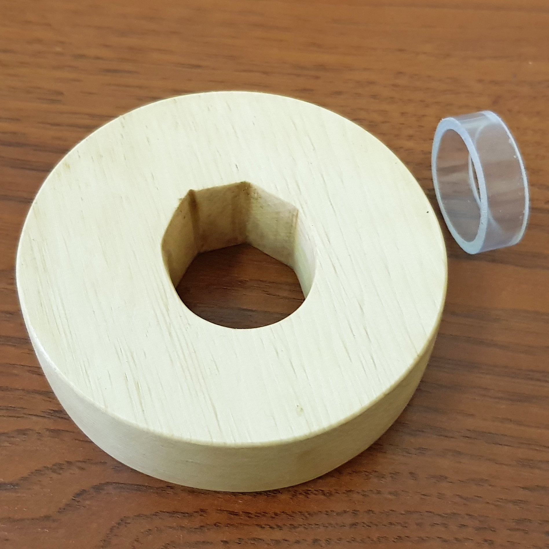 A wooden tsuba, which is a type of protective cover, placed next to a clear plastic clamp on a wooden surface.